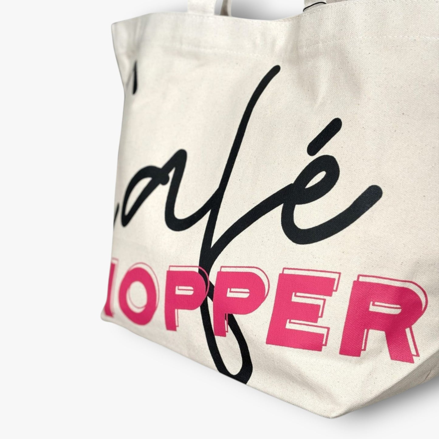 A canvas Café Hopper tote close-up showing bold black cursive and hot pink lettering.