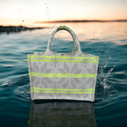 A tote bag with neon green stripes on light grey jacquard fabric on top of a pool of dark blue water. 