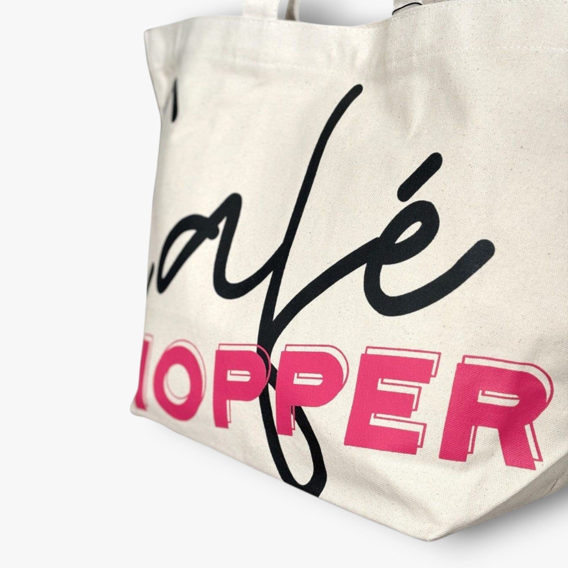 A canvas Café Hopper tote close-up showing bold black cursive and hot pink lettering.